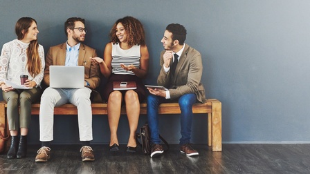Vier Personen sitzen auf einer Holzbank in einem Raum. Sie blicken sich gegenseitig an und interagieren miteinander. Auf ihren Schößen liegen von links nach rechts: Papiere, ein Laptop, ein Notizblock und ein Smartphone, ein Tablet