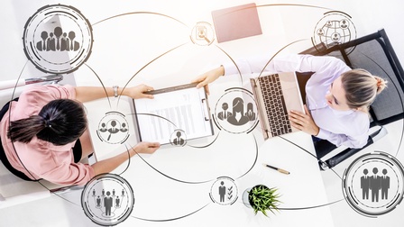 Zwei Personen sitzen sich an einem weißen Tisch gegenüber, die rechte tippt in einem Laptop, die linke hält ein Klemmbrett mit einem Dokument. Im Bild sind gezeichnete Linien, die zu Symbolen von Personen in Kreisen und einer Topfpflanze führen