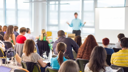 Rückenansicht vieler auf Sesseln sitzender Personen. Die Personen blicken zu einer stehenden Person, die beide Hände gehoben hat. Rechts hinter der Person ist eine weiße Tafel