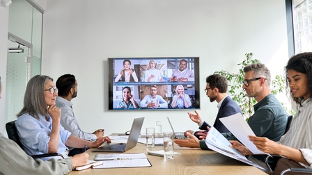 In einem Raum sitzen sich an einem Tisch Personen gegenüber. Insgesamt sind es sechs Personen. Auf dem Tisch sind Zettel, Laptops und Wasser Gläser. An der Wand hängt ein Bildschirm, auf dem sechs Personen sind