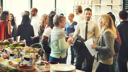 Mehrere Personen in Bürokleidung stehen in einem Raum in kleinen Gruppen und unteragieren miteinander. In der linken Bildhälfte ist ein gedeckter Tisch mit Gefäßen, befüllt mit mehreren Speisen. Im Hintergrund hängt eine dunkle, beschriftete Tafel
