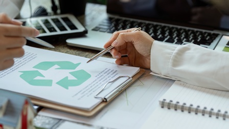 Auf einem Tisch liegt ein Klemmbrett mit einem Zettel, auf dem das Recyclingsymbol ist. Zwei Personen deuten jeweils mit einem Stift auf das Symbol
