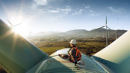 Person mit Helm und Sicherheitsgurten sitzt auf Turbine eines Windrades und blickt in hügelige grüne Landschaft mit weiteren Windrädern