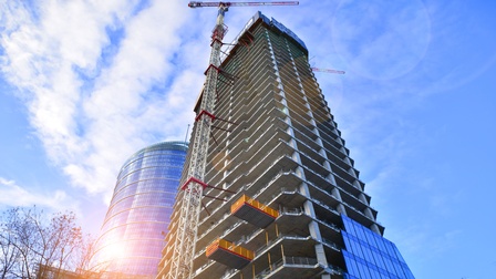 Ein Rohbau von einem Gebäude mit vielen Stockwerken unter blauem Himmel mit ein paar Wolken. Direkt neben dem Gebäude steht ein Kran.