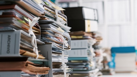 In der linken Bildhälfte sind viele Reihen übereinander gestapelter Aktenordner. In den Ordnern sind jeweils viele Zettel und Unterlagen