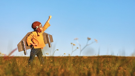 Ein Kind mit einer Fliegermütze und einer Fliegerbrille steht auf einer Wiese und streckt den linken Arm nach oben. Das Kind hat am Rücken gebastelte Flügel. Der Himmel ist blau