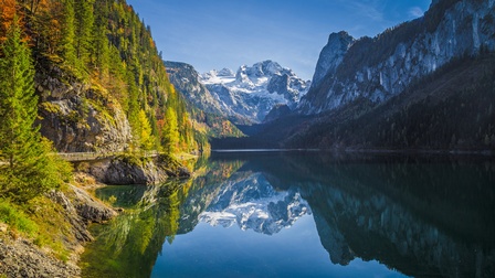 In einem Gewässer spiegelt sich hinten ein schneebedecktes Gebirge. In der rechten Bildhälfte spiegelt sich ein Gebirge, das im Schatten ist. In der linken Bildhälfte spiegelt sich ein Gebirge, das in der Sonne liegt