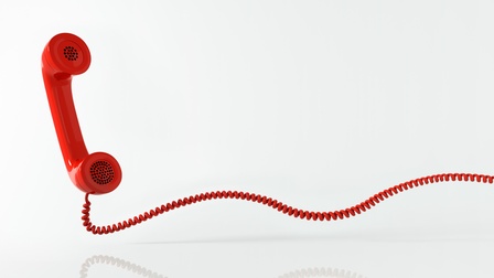 Nahaufnahme eines roten Schnur-Telefons vor weißem Hintergrund. In der linken Bildhälfte ist der Telefonhörer, die Schnur des Telefons reicht bis in die rechte Bildhälfte. Das Telefon ist auf glänzendem Untergrund platziert, wodurch ein Schatten entsteht