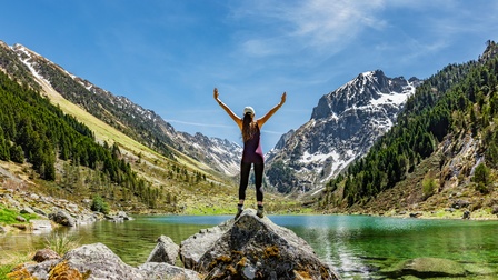 Person in sportlicher Kleidung mit langen dunklen Haaren steht auf einem Stein bei einem Bergsee und streckt die Hände Richtung Himmel