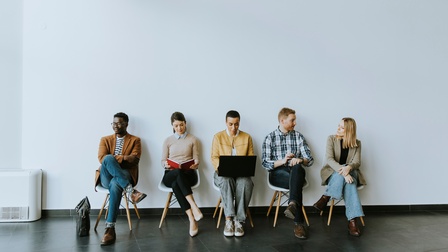 Vor einer hellen Wand sitzen auf fünf Sesseln nebeneinander fünf Personen. Eine Person blickt in ein Buch, eine auf einen aufgeklappten Laptop, zwei Personen unterhalten sich miteinander
