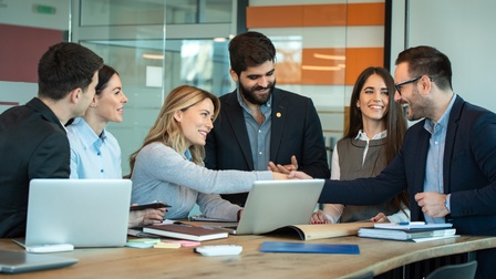 Mehrere lächelnde Person um Tisch versammelt, auf dem aufgeklappte Laptops stehen und Arbeitsutensilien liegen; zwei Personen reichen sich die Hand