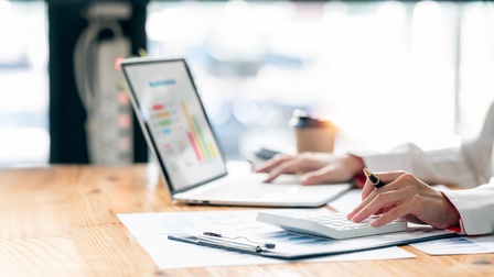 Seitliche Aufnahme einer Person, die an einem Tisch sitzt. Mit der linken Hand tippt sie auf einem Taschenrechner. Mit der rechten Hand bedient sie einen aufgeklappten Laptop, auf dem verschiedene Diagramme sind