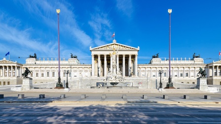 Außenansicht des österreichischen Parlamentgebäudes unter blauem Himmel: Grauweißer historischer Bau mit zentralem Säuleneingang, davor große Statue der Pallas Athene, ringsum und auf Dächern Figuren von Reitern