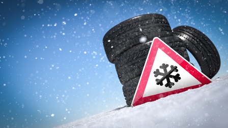 Illustration eines auf einem schneebedeckten Boden platzierten Dreiecks mit rotem Rand und dem Symbol einer schwarzen Schneeflocke. Dahinter sind Autoreifen übereinander gestapelt. Der Hintergrund ist blau, es sind Schneeflocken als Punkte angedeutet. 
