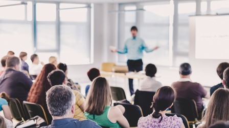 Rückenansicht vieler Personen, die auf Sesseln sitzen und zu einer Person blicken, die ihnen gegenüber steht und spricht. Vor der Person steht ein Tisch. Rechts neben der Person ist eine Leinwand