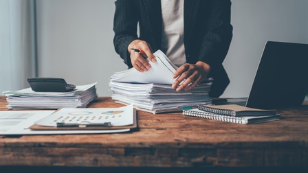 Eine Person in einem Sakko steht hinter einem Tisch. Vor ihr ist ein Papierstapel, den die Person durchsucht. Auf dem Tisch ist ein weiterer Papierstapel, ein Laptop, Notizbücher, ein Klemmbrett sowie ein Taschenrechner