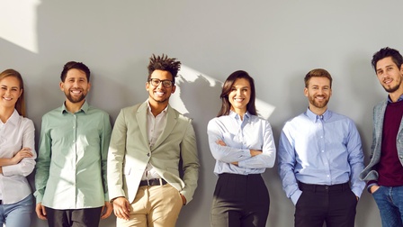 Mehrere lächelnde Personen stehen vor einer weißen Wand und blicken in die Kamera, während Sonnenlicht seitlich hereinstrahlt