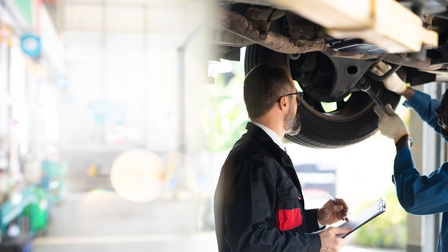 Zwei Personen stehen nebeneinander unter einem erhöhten Fahrzeug, das auf einer Hebebühne steht. Die rechte Person hält eine Zange. Die linke Person hält ein Klemmbrett und einen Stift