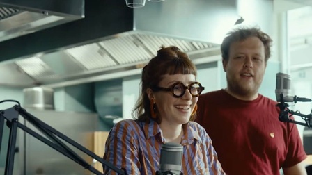 Frontale Aufnahme von zwei Personen, die hinter einem Tresen stehen. Vor den Personen sind zwei Mikrofone angebracht. Hinter ihnen ist eine Küche