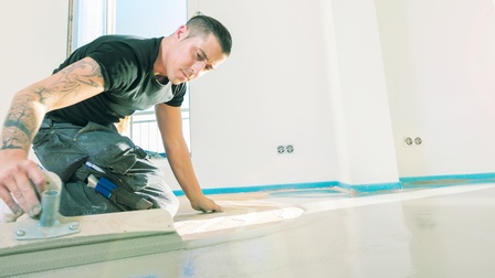 Eine Person kniet auf dem unverputzten Boden eines hellen, großen Raums und verteilt darauf eine Schicht Beton