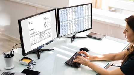 Person sitzt an Schreibtisch vor Computermonitoren mit Tabellen und Rechnung und tippt auf Tastatur