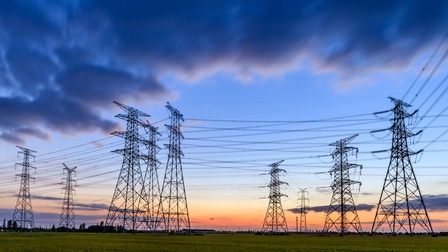 Mehrere Strommasten stehen auf einer flachen, grünen Landschaft. Zahlreiche Hochspannungsleitungen verlaufen zwischen den Masten über die gesamte Bildbreite