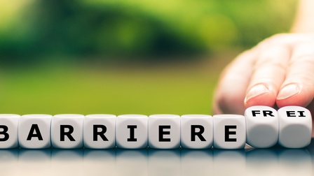 Weiße Holzwürfel nebeneinander stehend mit schwarzer Aufschrift barrierefrei, die letzten beiden Würfel werden von einer Hand nach hinten gekippt mit dem Wort frei