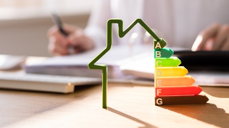 Energieeffizienz-Haus mit Stufen im Fokus, im Hintergrund befindet sich eine Person und sichtet Unterlagen während ein Stift und ein Rechner bereit stehen