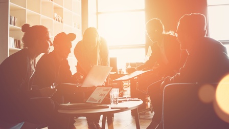 Gruppe von Personen sitzt rund um einen Tisch mit Arbeitsunterlagen und Laptops zusammen. Die Sonne scheint so durch das Fenster, dass man nur die Silhouetten der Personen sieht