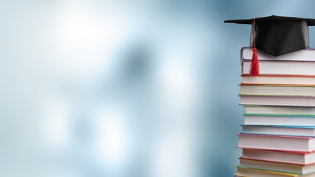 Ein hoher Bücherstapel steht vor einem hellgrauen, verschwommenen Hintergrund. Auf dem Stapel liegt eine abgeflachte, schwarze Kappe