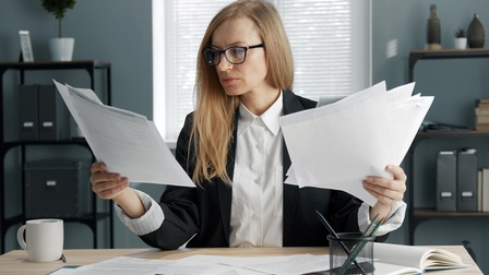 Person mit Brillen sitzt an Tisch und sichtet Dokumente ,weitere Unterlagen und Tasse auf Tisch platziert, im Hintergrund verschwommen Regale mit Ordnern