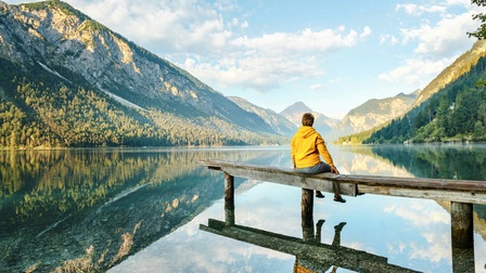 Person in gelbem Pullover sitzt auf in See reichenden Holzsteg umgeben von Berglandschaft, die sich in See spiegelt