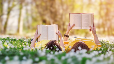 Zwei Personen liegen in Blumenwiese und lesen Bücher, im Hintergrund Wald