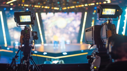 Zwei verschwommene Köpfe von Personen, die Hinter Filmkameras stehen, die Kameras auf große Showbühne gerichtet