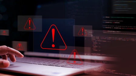 Eine Person hat ihre linke Hand bei der Tastatur eines aufgeklappten Laptops. Über der Tastatur sind sechs rote Dreiecke, in denen jeweils ein Rufzeichen ist