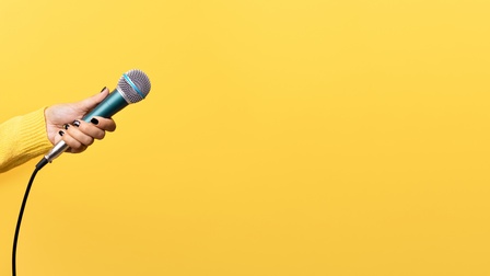 Vor einem gelben Hintergrund hält die linke Hand einer Person ein Mikrofon mit Kabel ins Bild. Die Person trägt ein gelbes Oberteil und hat lackierte Fingernägel