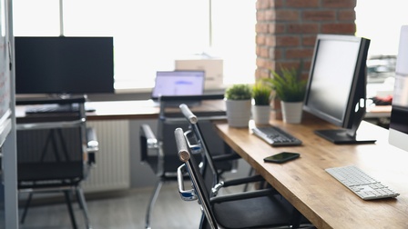 In der rechten Bildhälfte steht ein Schreibtisch mit zwei Bildschirmen, zwei Tastaturen, zwei Stühlen. Im linken Bildrand steht ein beschriftetes Whiteboard. Im Hintergrund stehen weitere Büromöbel und Büroutensilien.