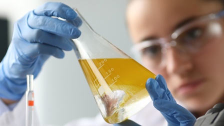 Im Fokus Laborglas mit gelber Flüssigkeit, das Person mit Schutzbrillen verschwommen im Hintergrund neigt und darauf blickt