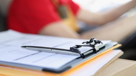 <br />
Detailaufnahme von einem Klemmbrett mit Unterlagen und Stift, im Hintergrund sitzt verschwommen eine Person am Steuer eines Fahrzeuges