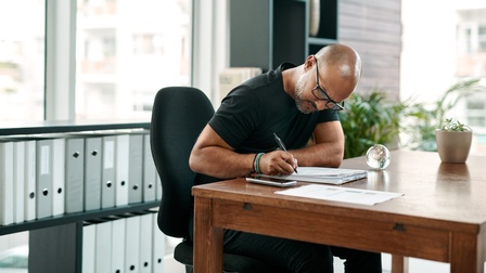 Eine schwarz gekleidete Person mit Brille sitzt in einem Büro und schreibt fokussiert.