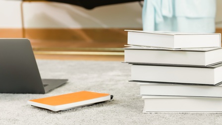 Ein aufgeklappter Laptop und ein Bücherstapel liegen auf einem hellen Teppich am Boden.