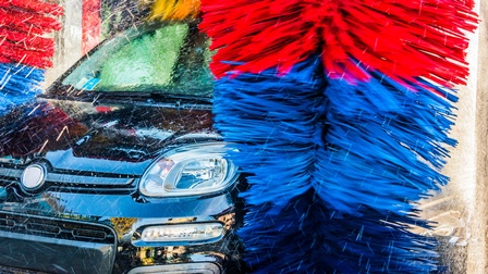 Zwei große Quasten in den Farben blau und rot stehen links und rechts von einem schwarzen Auto, das Auto ist nass, Wassertropfen fliegen durch die Luft.