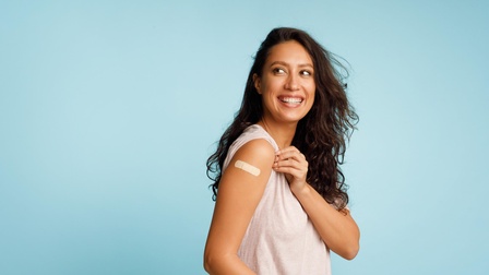 Eine junge Frau zeigt lächelnd ihr Pflaster von einer Impfung auf dem Oberarm