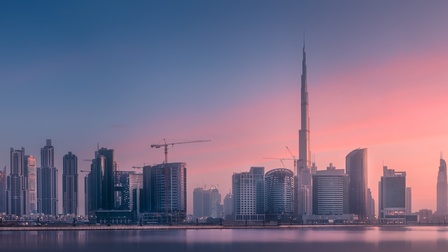 Panorama einer modernen Skyline mit zahlreichen Hochhäusern entlang eines Gewässers. In der Bildmitte ragt ein besonders hoher Turm über die übrigen Gebäude hinaus. Mehrere Baukräne sind zwischen den Gebäuden