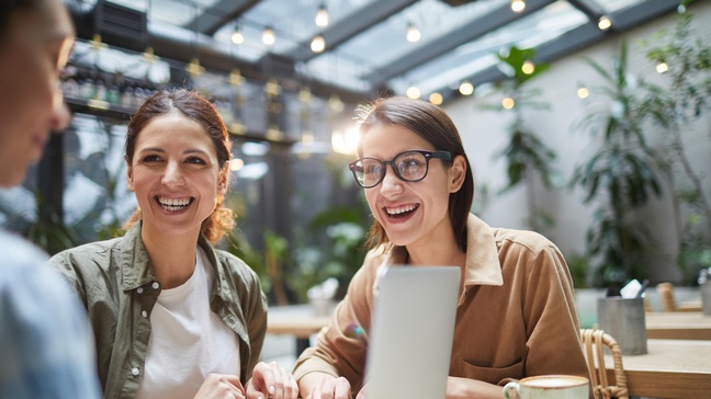 Zwei lächelnde Personen sitzen am Tisch mit weiterer Person vor Laptop, Kaffeetassen am Tisch, im Hintergrund Lichterketten und Pflanzen
