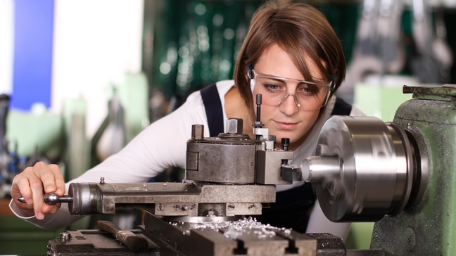 Person mit Schutzbrillen bedient Drehmaschine, in der Metallstück geschliffen wird und blickt auf Werkstück