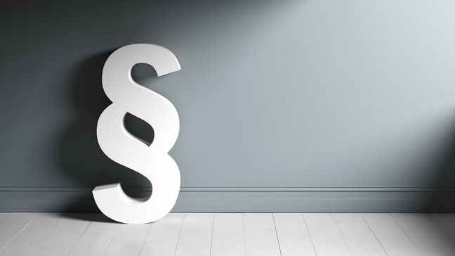 Weißes Paragrafenzeichen lehnt an grauer Wand auf hellem Parkettboden stehend