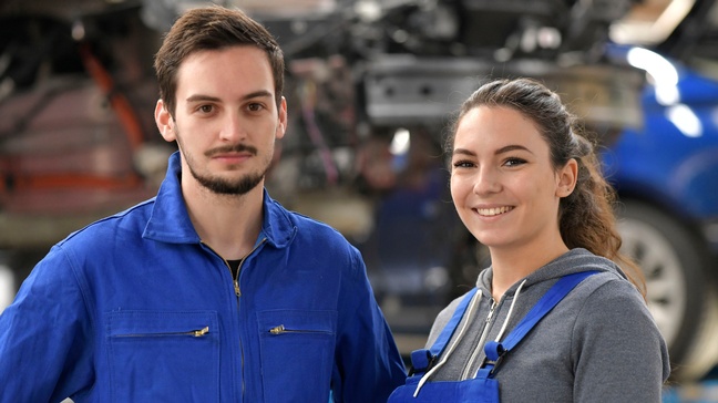 Zwei lächelnde Personen in blauer Arbeitskleidung, im Hintergrund verschwommen ein Auto