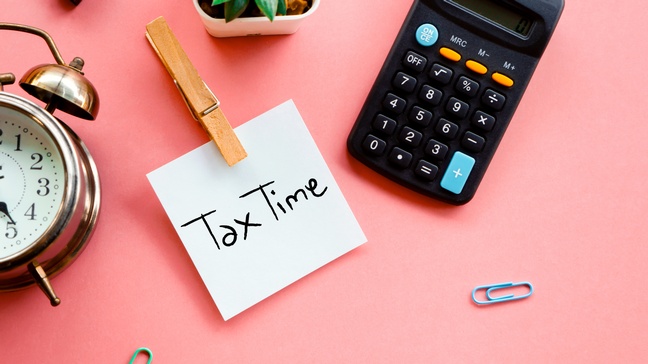Bildkonzept mit Post-It Tax Time, daneben liegen ein Taschenrechner, eine Zierpflanze, ein Wecker sowie Büroklammern auf einem rosaroten Hintergrund