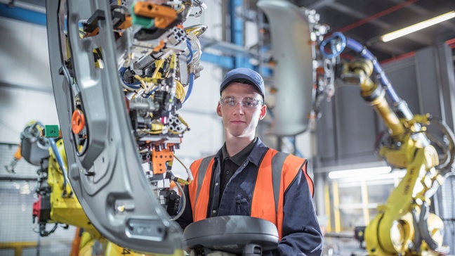 Portrait einer Person in Arbeitskleidung mit Kappe und Schutzbrille steht neben einem Roboterarm in einer Werkstätte